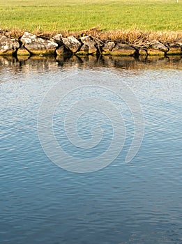 Vertical view of a river canal with deep blue water and a rocky edge and green grass behind abstract view