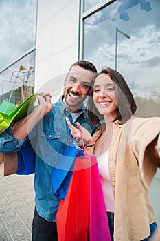 Vertical. Young caucasian real couple smiling and having fun taking a selfie portrait holding shopping bags at city