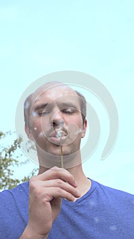 Vertically. young balding man blows on a dandelion on a background of blue sky