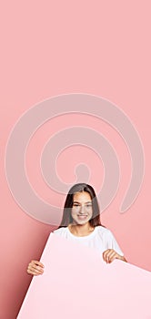 Vertival flyer with young pretty girl in white t-shirt holding huge empty sheet of paper isolated on pink background