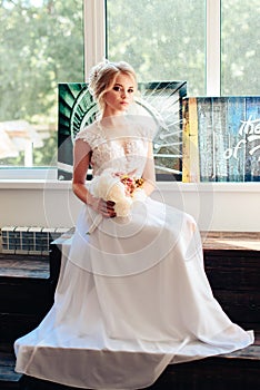 Very beautiful blonde with blue eyes in a white bride dress near a window with a bouquet of flowers