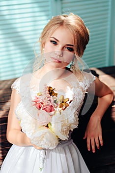 Very beautiful blonde with blue eyes in a white bride dress near a window with a bouquet of flowers