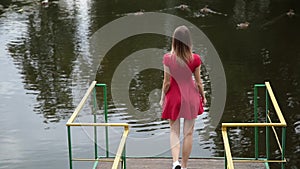 A very beautiful girl in a red dress is standing on a pier near a small lake.