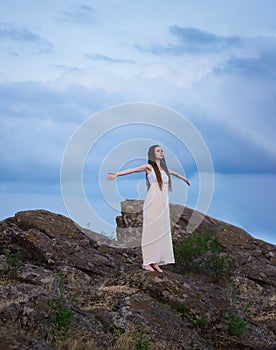 A beautiful girl in a white dress with dreadlocks is standing on a cliff with her arms outstretched against a cloudy sky at sunset