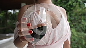 A very beautiful young girl in a soft pink evening dress with a large neckline and a red manicure shakes wine in a glass