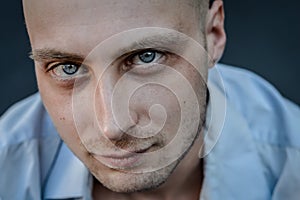 Very close portrait of a young bald man with blue eyes and a blue shirt looking intently at the camera