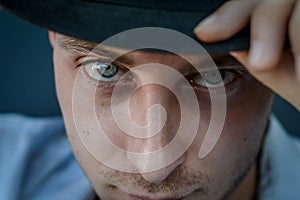 Very close portrait of a young blue-eyed man who with his hand adjusts the black hat on his head