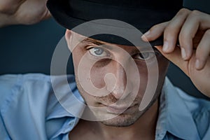 Very close portrait of a young blue-eyed man who with his hand adjusts the black hat on his head