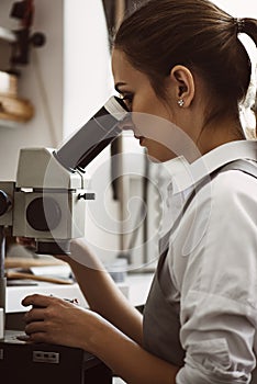 Very close. Vertical portrait of female jeweler looking at the new jewelery product through microscope in a workshop.