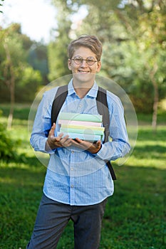 Very cute, young boy in round glasses and blue shirt with backpack holds books in the park. Education, back to school