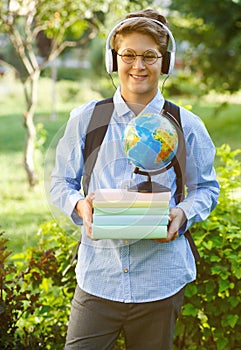 Very cute, young boy in round glasses and headphhones and blue shirt with backpack holds books, globe in the park. Education