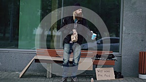 Very drunk homeless man with cardboard and alcohol sign beg for money while standing near bench at the sidewalk