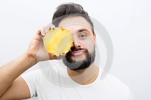 Very funny picture of positive young man stands and poses on camera. He coveres one eye with piece of pineapple. Young