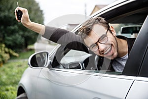 Very happy young man is peeking from the car window while looking at the camera. He is holding the keys at his right hand. Lottery