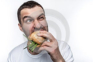 Very hungry man is eating a burger. He is biting it quite hard. Also man is looking somewhere aside. Isolated on white