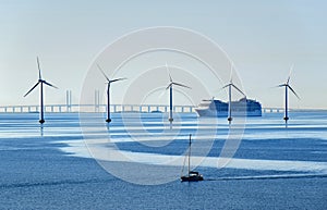 A very large passenger ship and a small sailboat pass offshore wind turbines near the Oresund Bridge between Denmark and Sweden