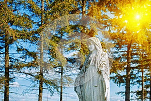 Very old and ancient stone statue of Virgin Mary in sunshine against blue sky and trees