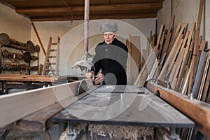 Very old man carpenter cutting wooden plank with circular saw in his workshop