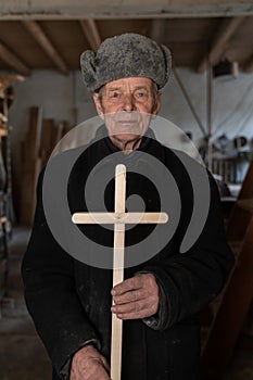 Very old man in retro clothes stands among his workshop at home, holds wooden cross in his hands and looking at the