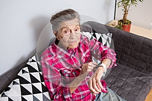 A very old senior Caucasian grandmother with gray hair and deep wrinkles sits at home on the couch in jeans and a red