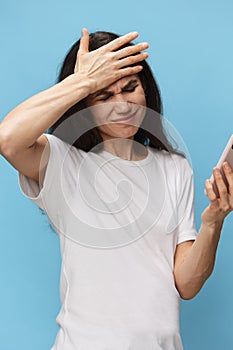 A very pensive woman stands on a blue background in a white t-shirt with a fashionable phone in her hands and looks at