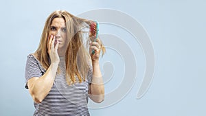 Very sad and upset young woman looking with shock at her damaged hair on blue background