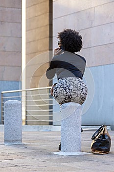 Very sexy Afro woman sitting on a stake in town is on the phone looking away. On the ground is her purse