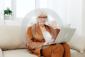 a very surprised elderly woman is sitting looking at a laptop on a sofa in a bright room near the window with glasses on