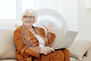 a very surprised elderly woman is sitting looking at a laptop on a sofa in a bright room near the window with glasses on