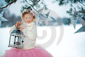 Very sweet beautiful little smiling girl child in pink skirt, white pullover, gray scarf and floral wreath with lantern in hands.