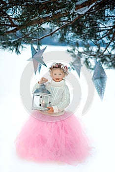 Very sweet beautiful little smiling girl child in pink skirt, white pullover, gray scarf and floral wreath with lantern in hands.