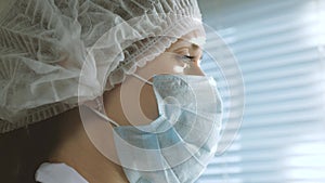 Very tired woman doctor in protective suit looks out window, takes off his medical mask, closing his eyes from fatigue