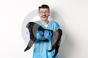 Vet clinic concept. Happy male doctor veterinarian holding cute black pug dog, smiling at camera, white background