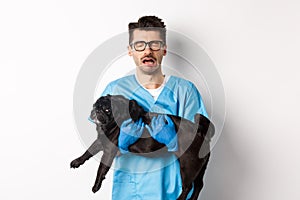 Vet clinic concept. Sad veterinarian holding black pug dog and crying, sobbing with miserable face, standing over white