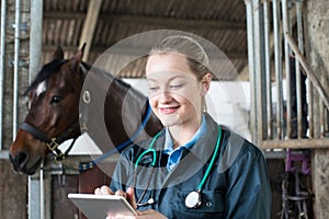 Female Vet With Digital Tablet Examining Horse In Stable