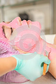 vHand of cosmetologist applying the pink alginic mask with the brush to the face of the young woman in a beauty salon.