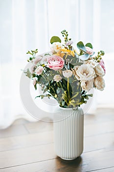 A vibrant bouquet of flowers in various colors and types, arranged in a white, ribbed vase on a wooden floor against the backdrop