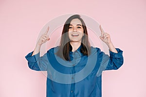 Vibrant Portrait of Energetic Young Woman Expressing Amazement with Open Mouth and Pointing Fingers Upward, Isolated on Pink