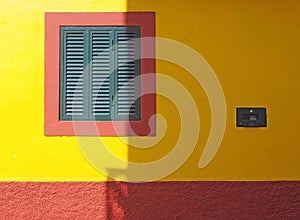 Vibrant red and yellow colorful house wall with a green wooden shutter in a square frame in bright sunlight and shadow