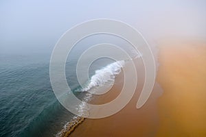 Vibrant Turquoise Ocean Wave Disappears into the Fog Along a High Contrast Orange Sandy Beach