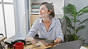 Vibrant young hispanic woman enjoying success at work, smiling while using touchpad in the busy office workspace, sipping coffee