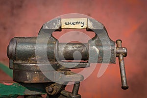 Vice grip tool squeezing a plank with the word enemy