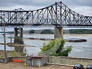 The Vicksburg Bridge over the flowing waters of the Mississippi river with a barge sailing underneath, lush green trees