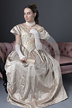 A Victorian woman wearing a gold evening gown and sitting on an antique sofa in front of a grey backdrop