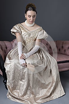 A Victorian woman wearing a gold evening gown and sitting on an antique sofa in front of a grey backdrop