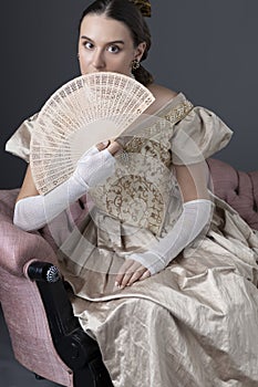 A Victorian woman wearing a gold evening gown and sitting on an antique sofa in front of a grey backdrop