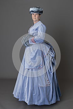 A Victorian woman wearing a pale blue velvet bustle ensemble with a fur muff and hat