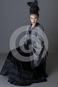 A Victorian woman wearing a striped silk polonaise and a black skirt and sitting down against a studio backdrop