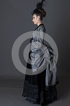 A Victorian woman wearing a striped silk polonaise and a black skirt and standing against a studio backdrop