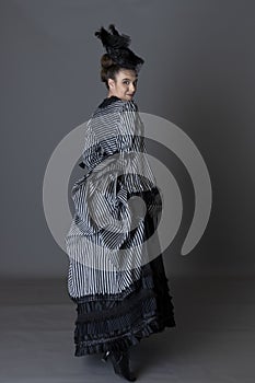 A Victorian woman wearing a striped silk polonaise and a black skirt and standing against a studio backdrop